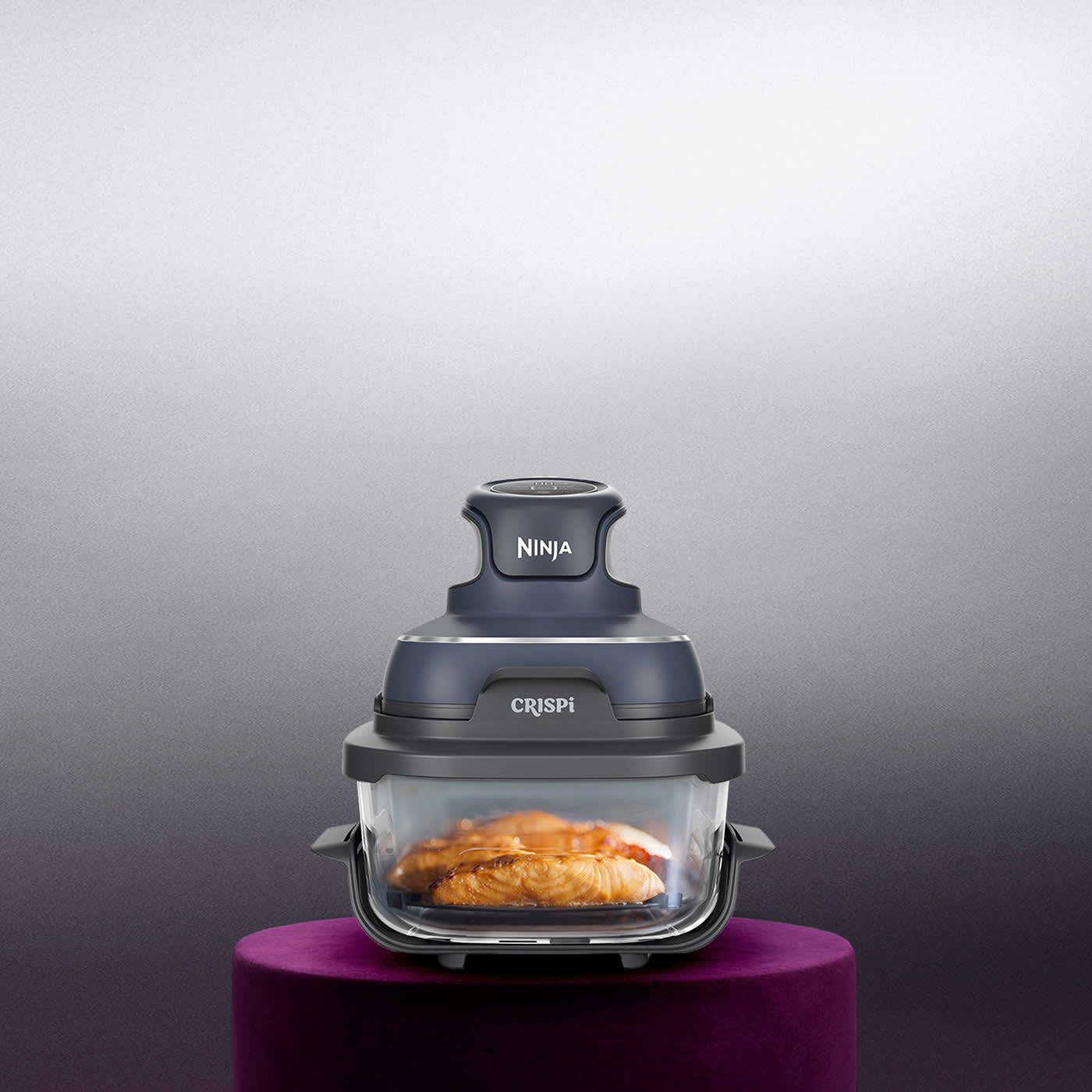 Ninja Crispi 4-in-1 portable glass air fryer with food inside, displayed on a purple pedestal against a grey background.